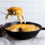 A hand uses a white-handled spatula to lift a portion of a melted cheese and shredded meat dish from a black cast iron skillet. A white plate and a red and white plaid towel are on a white marble countertop, with a white subway tile wall in the background.